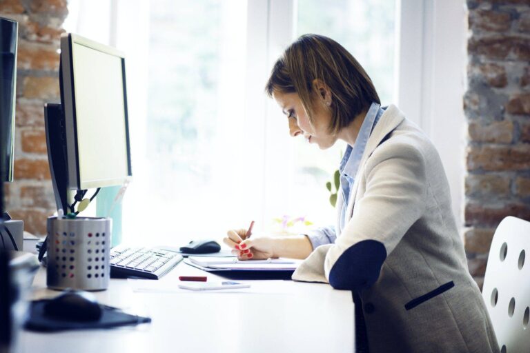 businesswoman writing in book by computer at desk in office