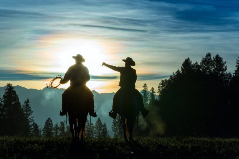 two cowboys riding across grassland with mountains in background early morning