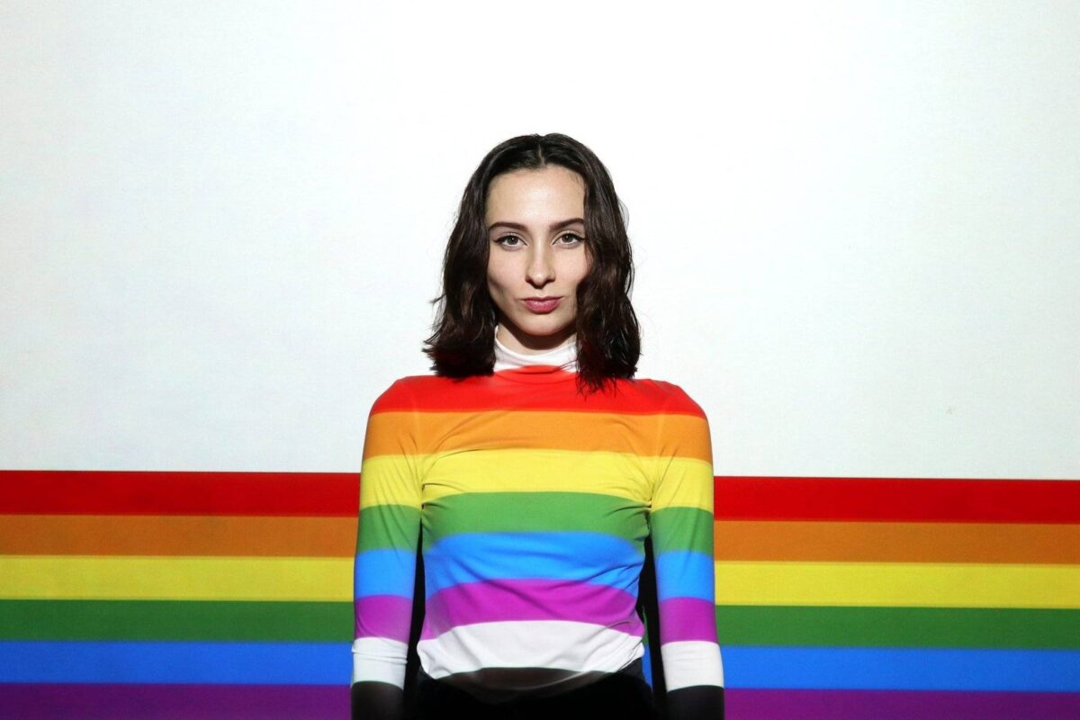 Eine Wand hat horizontale Regenbogen-Streifen. Eine Frau steht davor, ihr Pullover hat dieselben Regenbogen-Streifen wie die Wand.
