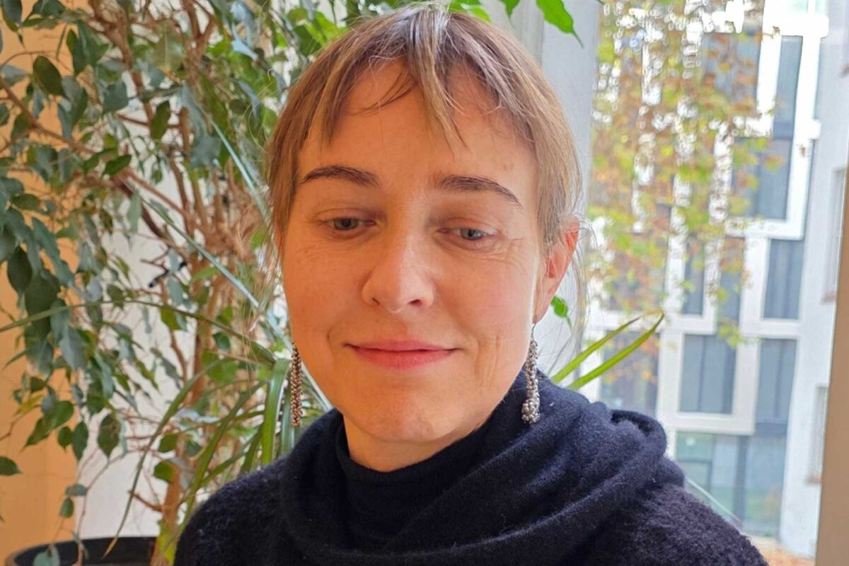 Porträt einer Frau mit kurzen Haaren und silbernen Ohrringen, sitzend vor einem Fenster mit Pflanzen im Hintergrund. Es handelt sich um Heike Sohna, die Geschäftsführerin von SEQUOYA.