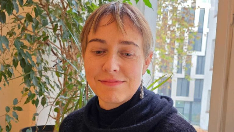 Porträt einer Frau mit kurzen Haaren und silbernen Ohrringen, sitzend vor einem Fenster mit Pflanzen im Hintergrund. Es handelt sich um Heike Sohna, die Geschäftsführerin von SEQUOYA.