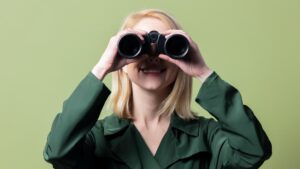 Blonde Frau in dunkelgrüner Bluse blickt lächelnd durch ein Fernglas vor hellgrünem Hintergrund.