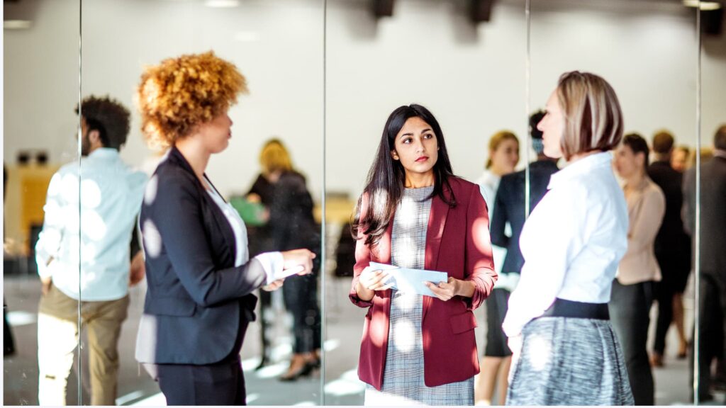 Drei Frauen stehen bei einer Networking-Veranstaltung in einem hellen Konferenzraum zusammen und unterhalten sich. Im Hintergrund sind weitere Teilnehmende unscharf zu sehen. Eine Frau in rotem Blazer hält Notizen in der Hand und hört aufmerksam zu.
