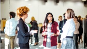 Drei Frauen stehen bei einer Networking-Veranstaltung in einem hellen Konferenzraum zusammen und unterhalten sich. Im Hintergrund sind weitere Teilnehmende unscharf zu sehen. Eine Frau in rotem Blazer hält Notizen in der Hand und hört aufmerksam zu.